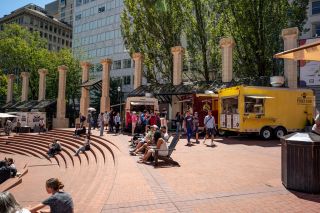 people sit in chairs and on steps in front of food carts in a brick courtyard in the center of an urban downtown area
