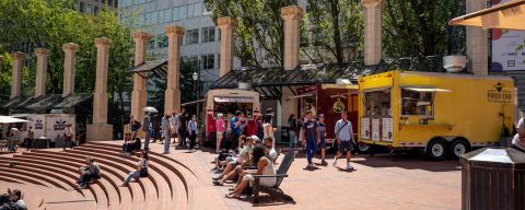 people sit in chairs and on steps in front of food carts in a brick courtyard in the center of an urban downtown area