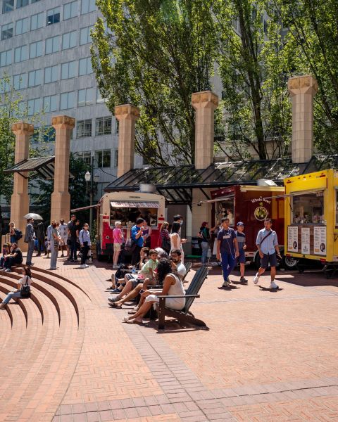 people sit in chairs and on steps in front of food carts in a brick courtyard in the center of an urban downtown area