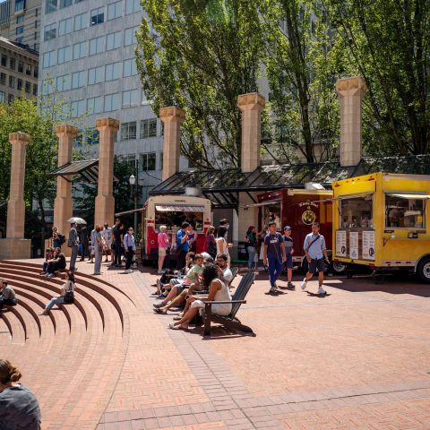 people sit in chairs and on steps in front of food carts in a brick courtyard in the center of an urban downtown area