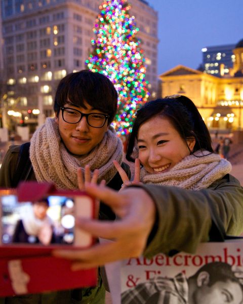 a couple poses for a selfie in front of a Christmas tree and a historic building