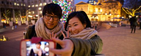 a couple poses for a selfie in front of a Christmas tree and a historic building