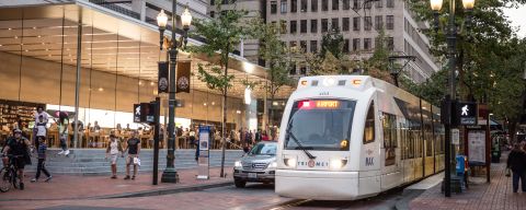 A light rail train passes in front of a modern, glass-encased retail store