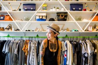 a woman with tattoos and wearing a hat stands in front of a rack of clothing at Wildfang clothing store