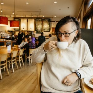 a white person with long hair on one side and a shaved head on the other side, sips from a coffee mug in a warmly lit bakery.
