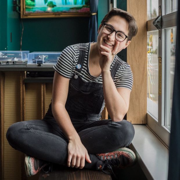 a smiling person sitting cross-legged on a wooden bench in front of a window