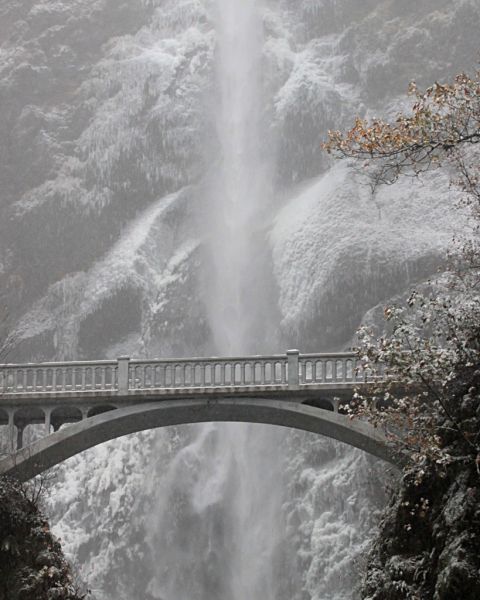 a towering waterfall behind a stone bridge surrounded by snow and ice