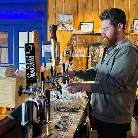 A man wearing a gray hoodie pours a glass of beer from a beer tap inside a wood-paneled bar.