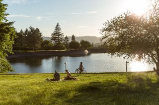 a biker and two picnicking people in front of a tree lined reservoir