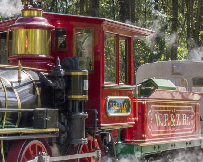 A bright red vintage steam train rounds a bend with deciduous forest int he background