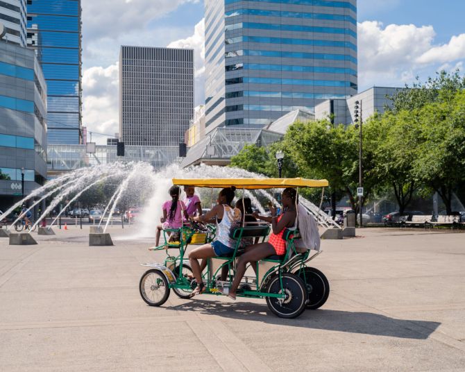 people ride a pedicab in front of a large fountain in an urban park on a sunny day