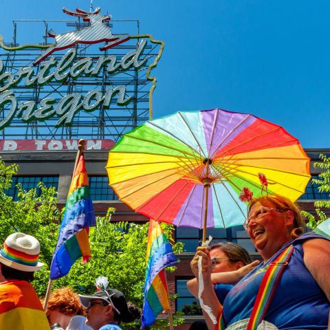 person with rainbow suspenders and rainbow parasol smiles, surrounded by people in rainbow decoration with Portland\'s white stag sign above