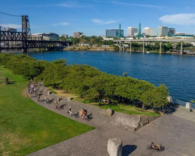 People ride bikes on a sunny day through a park along a river with bridges and city views in the background.