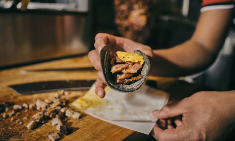 a close up image of hands serving a taco with blue corn tortillas, roasted meat and a piece of pineapple sitting on top