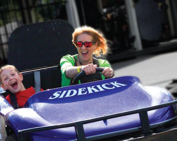 an adult and a child smile widely while racing along in a purple go-kart