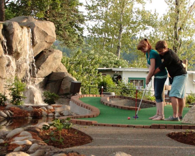 one person reaches around another to guide a short putt on a miniature golf course