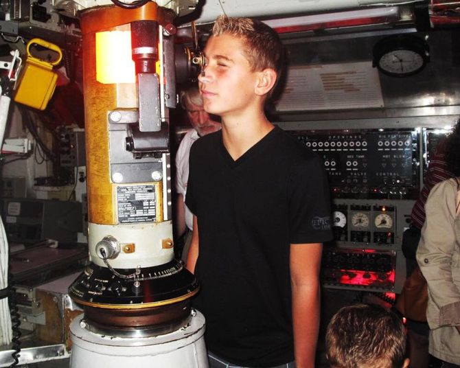 an adolescent boy in a submarine peers through a scope while other people look on
