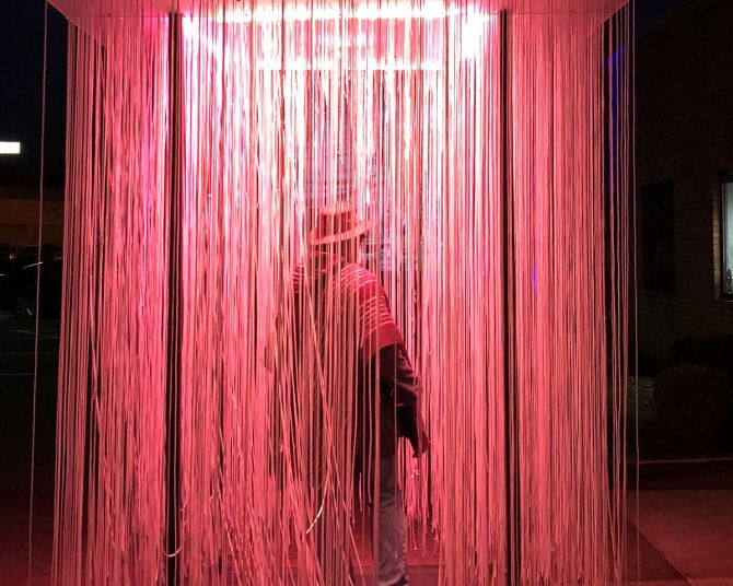 a person walks through a light installation containing long strands of lights hanging from a panel overhead