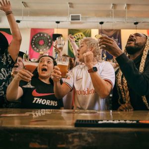 a crowd of people cheer while raising glasses in front of a bar while watching a game