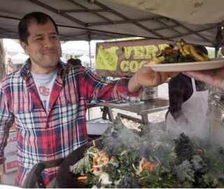 man in an outdoor market handing a plate of food to a customer in front of a sign reading "Verde Cocina"