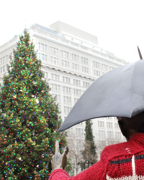 a large tree decorated with lights in front of a statue of a person holding an umbrella