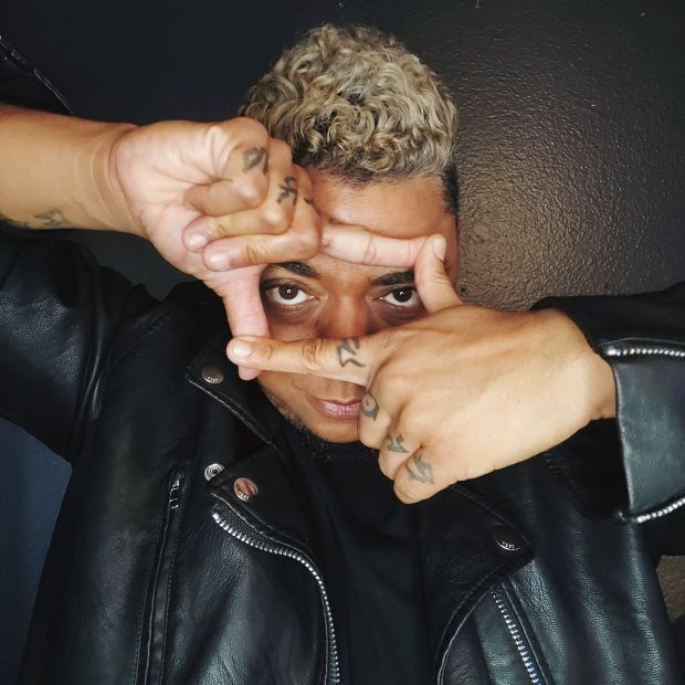 A Black man with blond hair and wearting a black leather jacket looks at the camera through a rectangle created with his own fingers held in front of his face