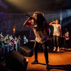 two hip hop performers sing into microphones on stage in front of an excited audience