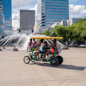 people ride a pedicab in front of a large fountain in an urban park on a sunny day