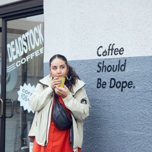 person stand outside of a store front with a coffee there is a mural on the building that says Coffee Should Be Dope