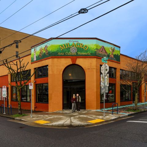 a couple stands in front of a large theater painted orange with a sign reading “En Centro Milagro”