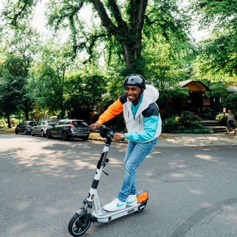 a smiling man wearing a helmet rides a Biketown scooter on a leafy city street
