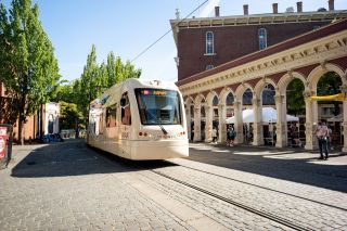 a light rail train on a cobblestone road passes the historic Saturday Market entry arches
