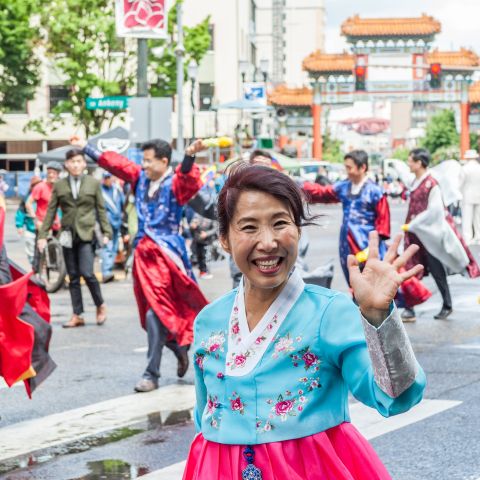 A woman wearing a hanbok and a big smile waves at the camera among a crowd of parade marchers in front of the ornate gate in Old Town China Town.