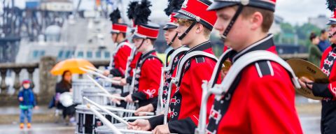 youth drum line of marching band in red costumes and hats walk along bridge in parade