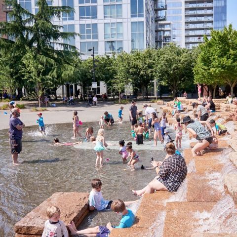 many children and a few adults play in the fountain at Jamison Square on a bright summer day