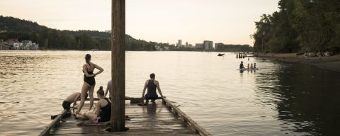 a wooden dock in a river — with swimmers standing, sitting or climbing out of the water — looks toward a city skyline in the fading sunlight