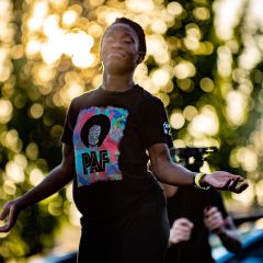 woman holds hands and arms out in dance performance in a park, sun filters through the leaves of the tree behind her