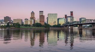 pale pink and purple sky reflect on a river surface with a bridge off to the side and a view of downtown in the background