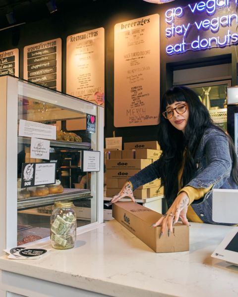 a woman at a counter closes the lid on a box of doughnuts at the counter of a bakery behind a glass cabinet full of doughnuts and desserts