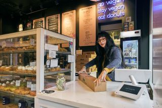 a woman at a counter closes the lid on a box of doughnuts at the counter of a bakery behind a glass cabinet full of doughnuts and desserts