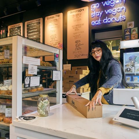 a woman at a counter closes the lid on a box of doughnuts at the counter of a bakery behind a glass cabinet full of doughnuts and desserts