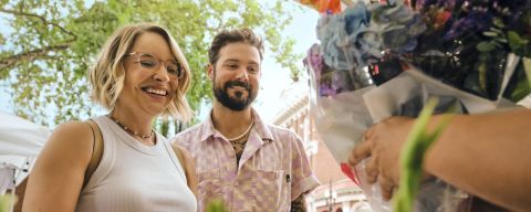 people smiling on a sunny day in front of bunches of flowers while someone off screen is handing them a bundle of cut flowers in a plastic bag