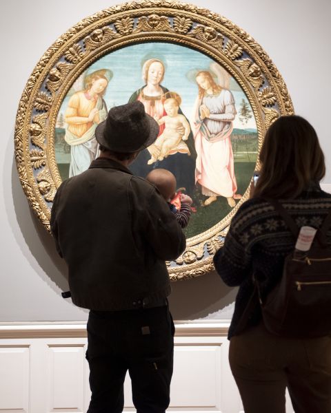 two people and a baby stand in front of a round oil classical painting of a religious scene at the Portland Art Museum