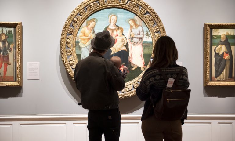 two people and a baby stand in front of a round oil classical painting of a religious scene at the Portland Art Museum