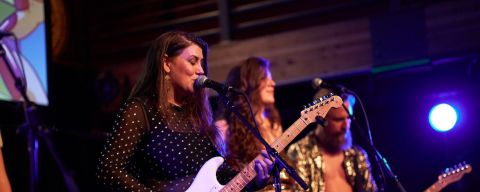 person sings in to a microphone while playing a guitar with two band members behind on a stage.