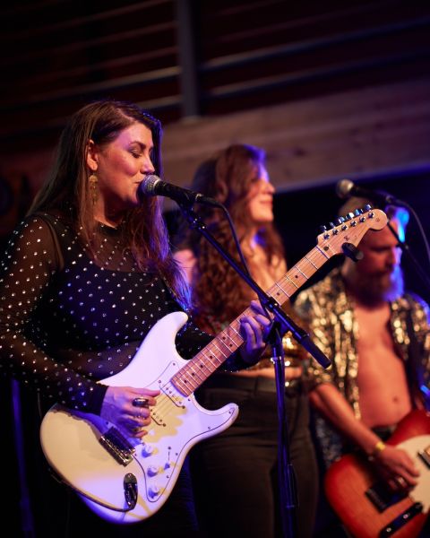 person sings in to a microphone while playing a guitar with two band members behind on a stage.