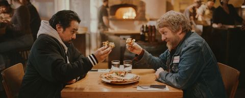 two men seated at a table share a pizza in a warmly lit pizzeria with a wood-fired oven in the background