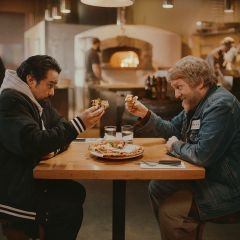 two men seated at a table share a pizza in a warmly lit pizzeria with a wood-fired oven in the background