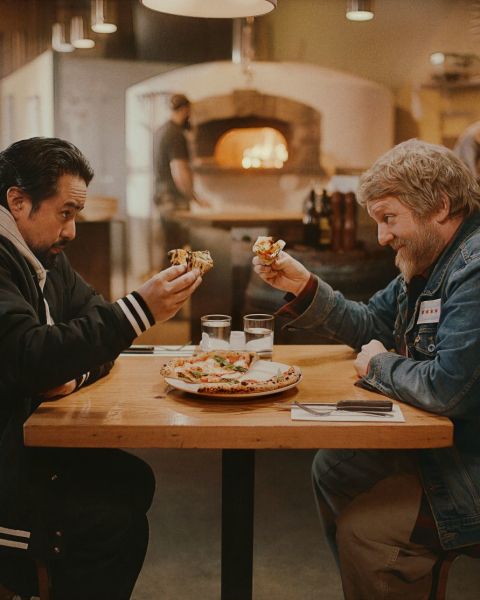 two men seated at a table share a pizza in a warmly lit pizzeria with a wood-fired oven in the background