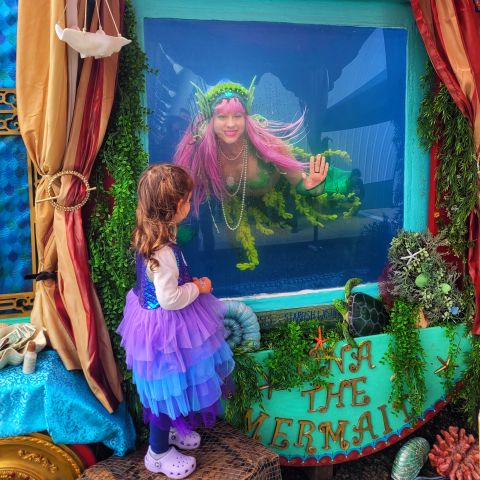 a person dressed as a mermaid swims inside a water tank as a child looks on